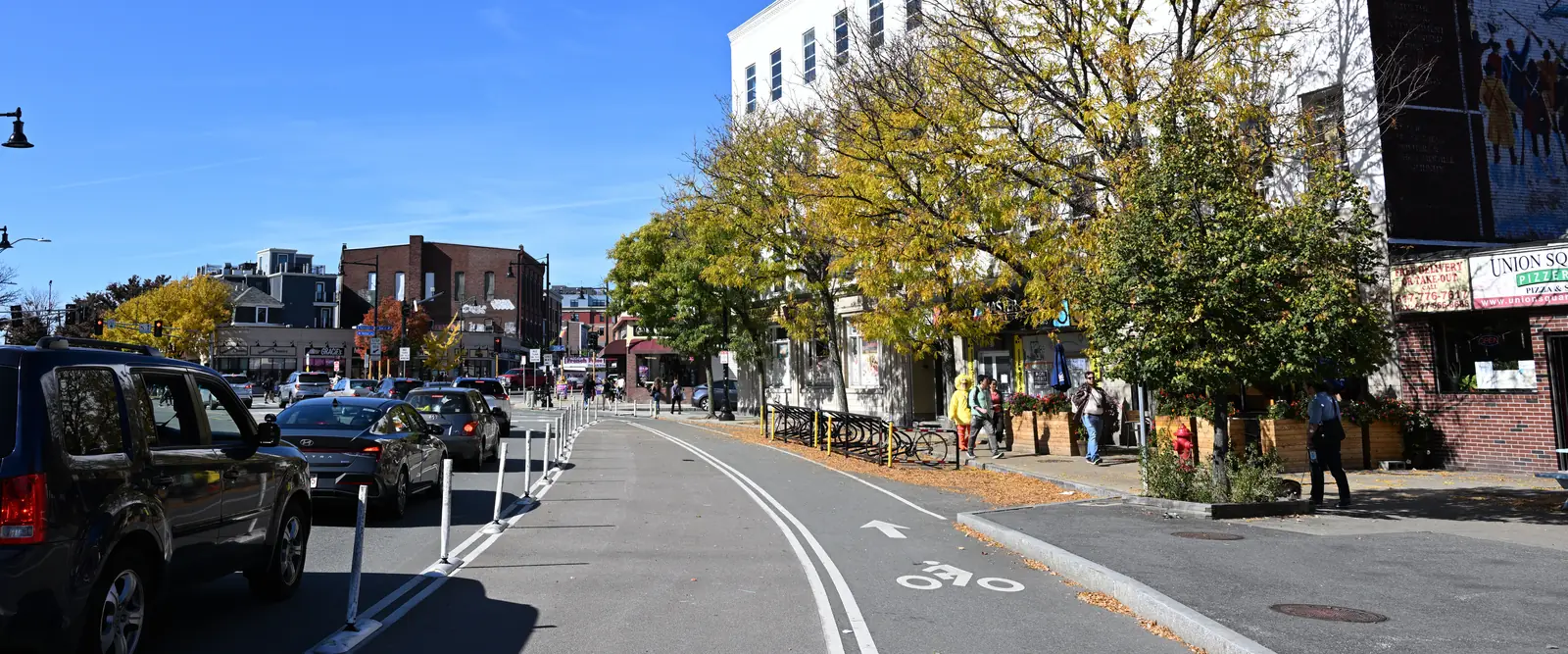 Street and storefronts in Union Square, Somerville, with autumn trees and a cycle lane